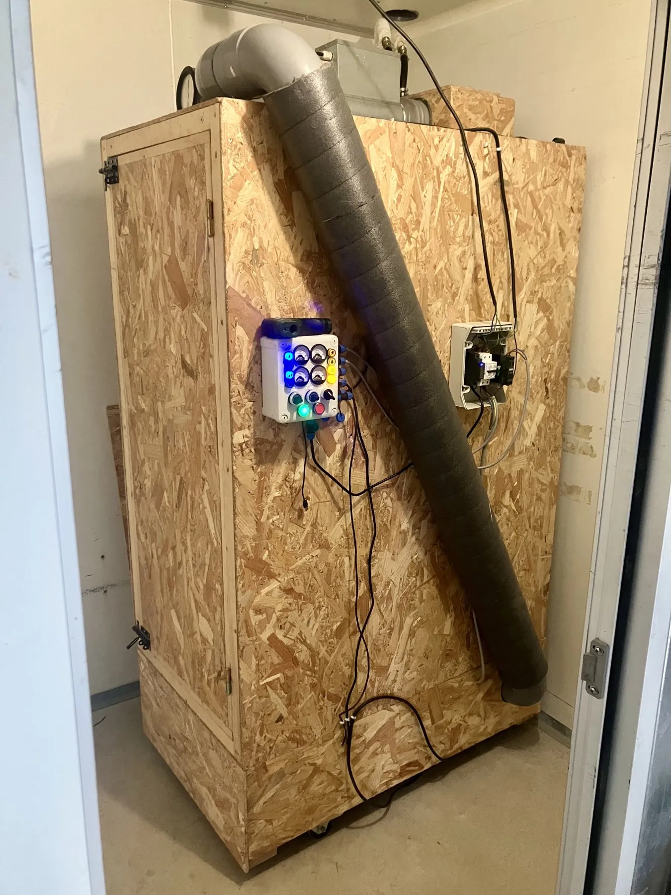 Le prototype en OSB dans son local : la porte à gauche, une gaine isolée en diagonale sur le côté et les coffrets de commande.